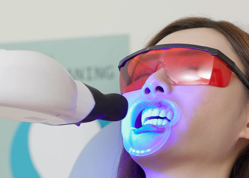 Dentist performing teeth whitening treatment on patient wearing protective orange goggles with blue LED light